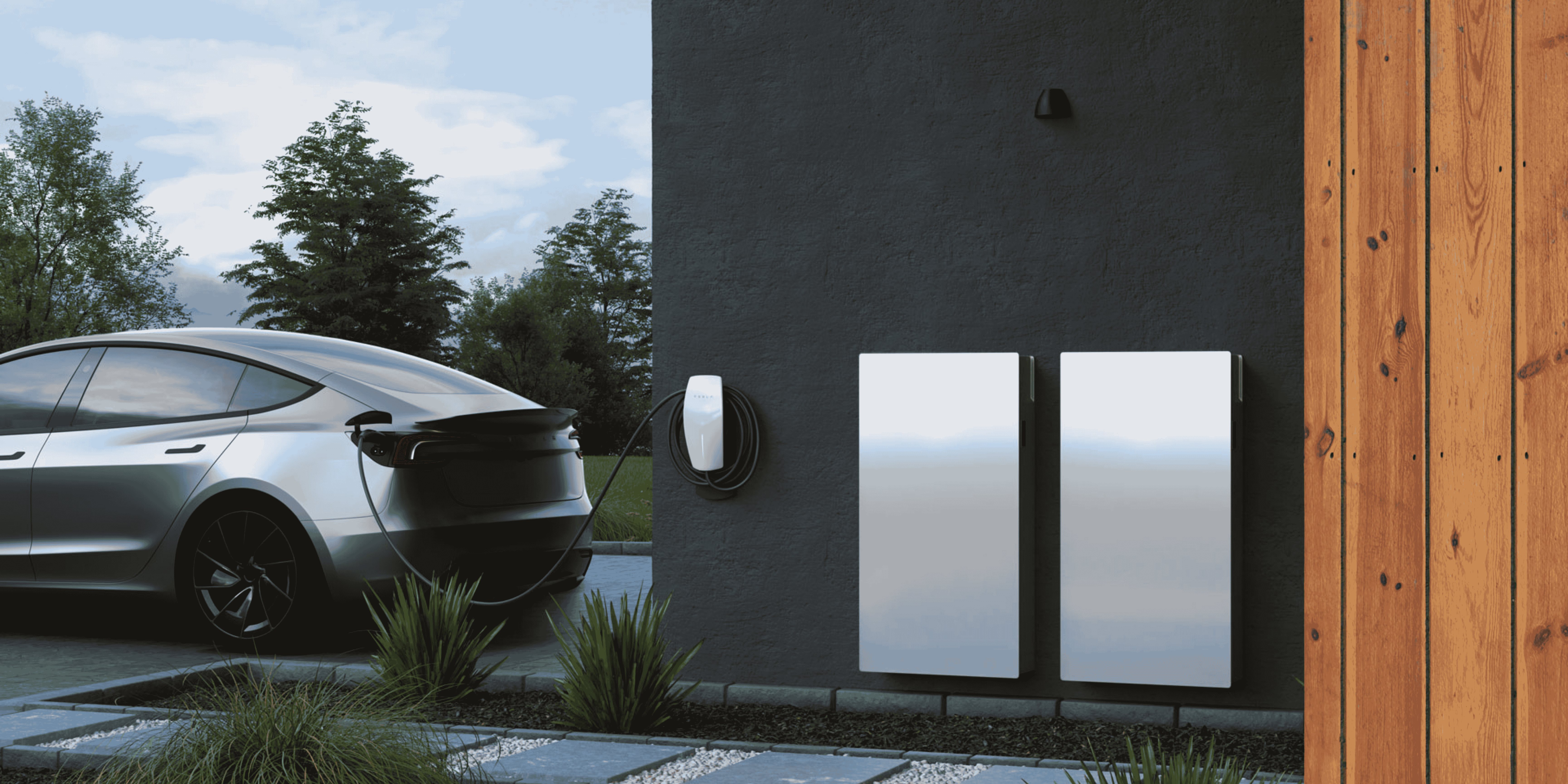 Silver electric car charging on a driveway next to a black wall with two mounted battery units.