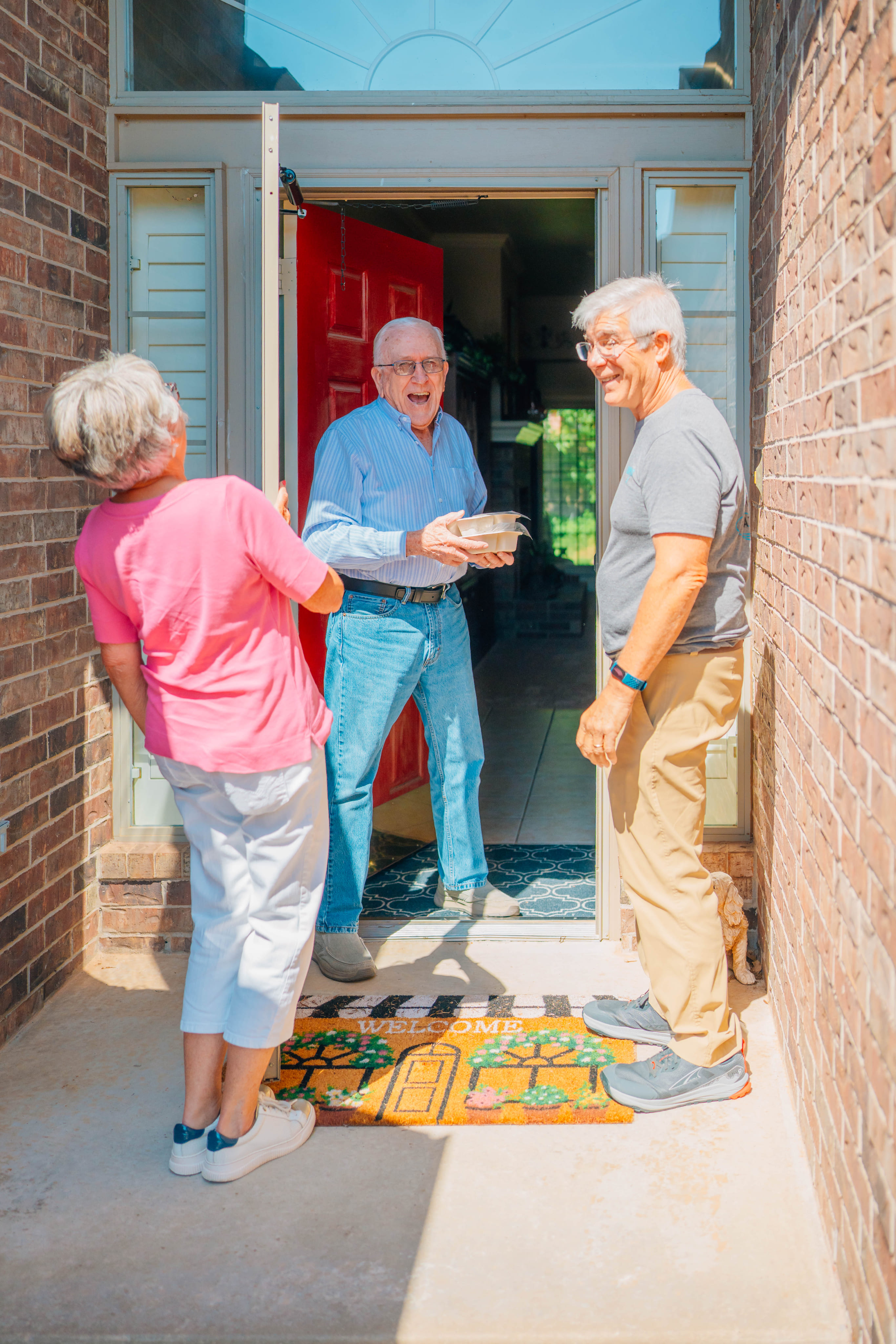 An older man stands in a doorway holding a tray while two older adults talk with him on a brick front entryway with colorful mats.
