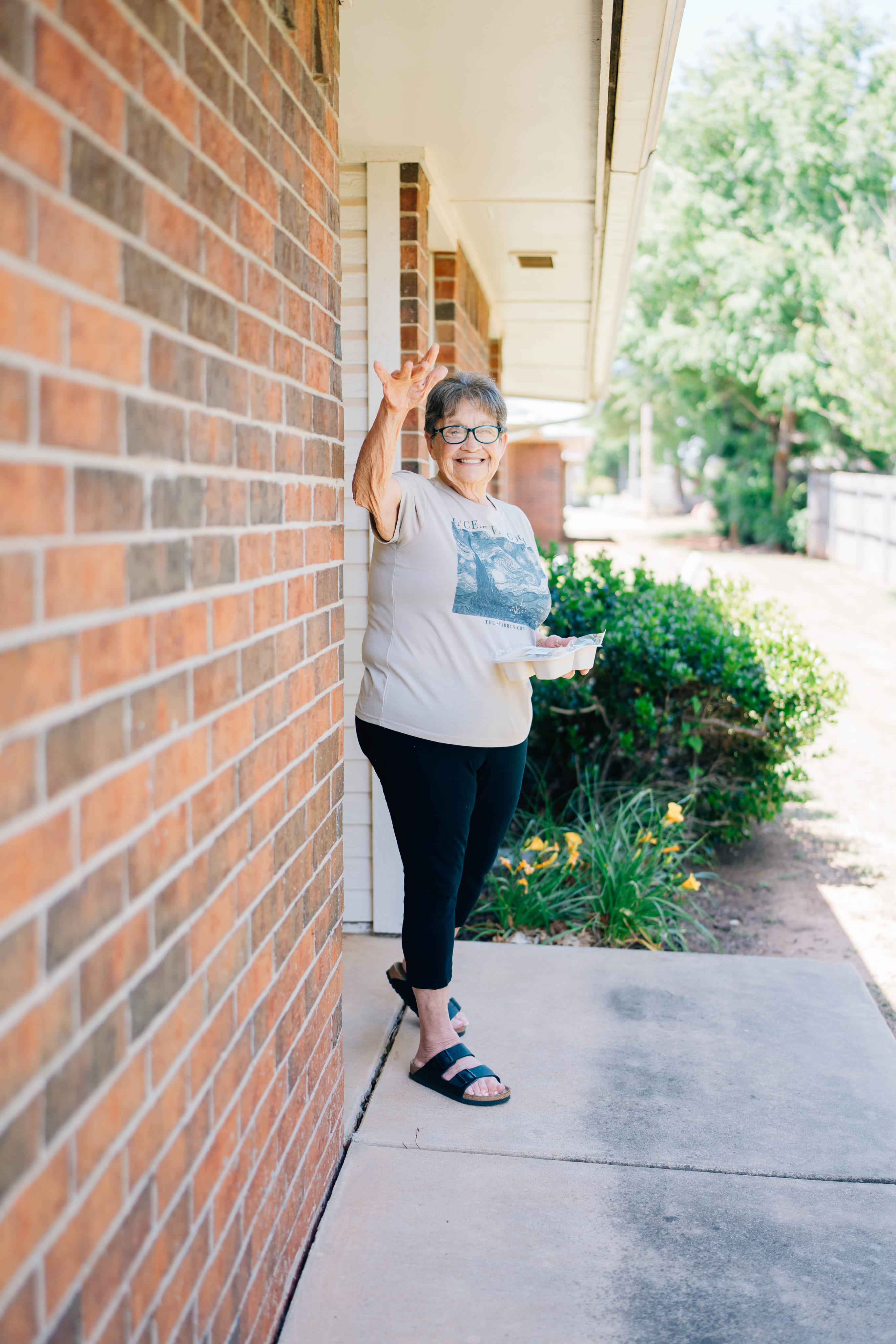A woman with glasses stands by a brick wall on a sidewalk, raising two fingers while holding a plastic cup, with bushes and trees in the background.