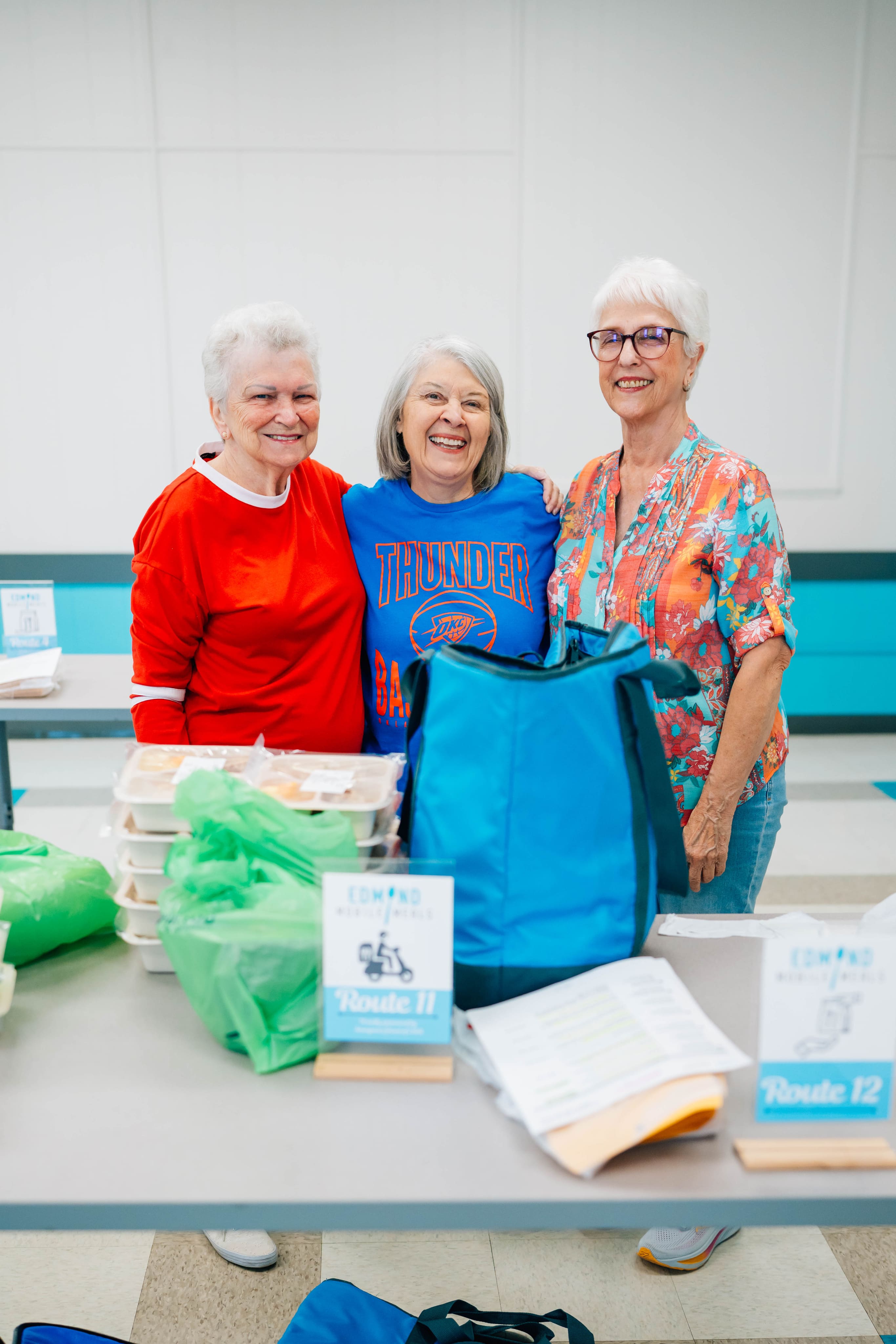 Three older women stand behind a table with blue and green reusable bags, brochures, and folded items in a bright indoor room.