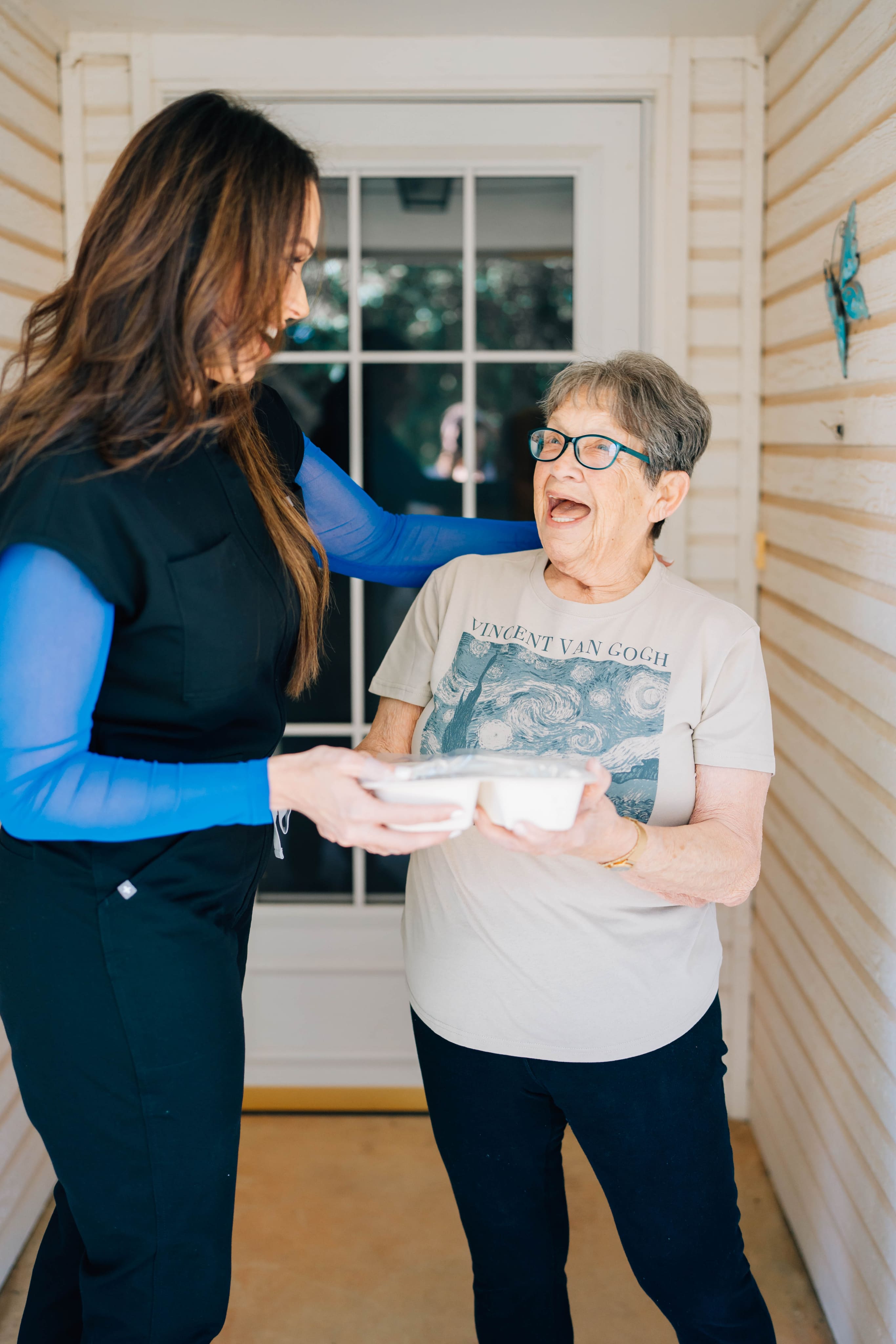A woman in a black vest and blue shirt hands a meal container to an older woman wearing glasses and a Vincent van Gogh T-shirt, standing in a doorway.