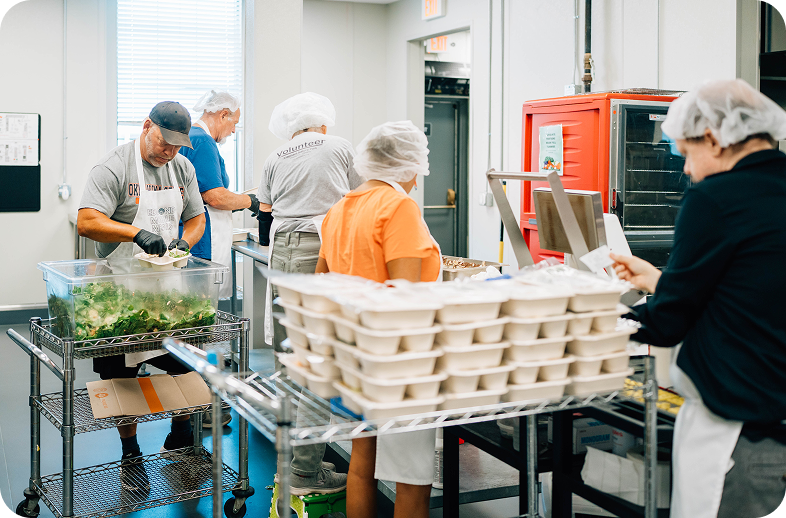 Workers in hairnets and aprons prepare food in a commercial kitchen, with stacked takeout containers on a metal cart and a red warming cabinet in the background.