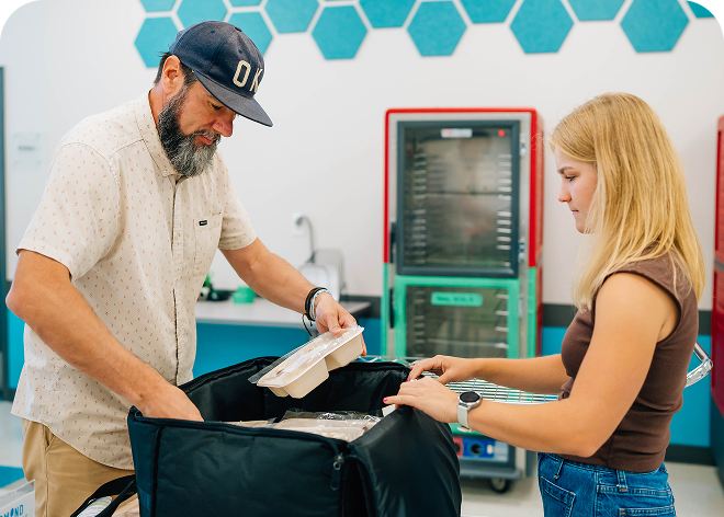 A bearded man in a cap and a woman stand beside an open black insulated bag, handling a foam takeout container in a room with teal counters and a glass refrigerator.