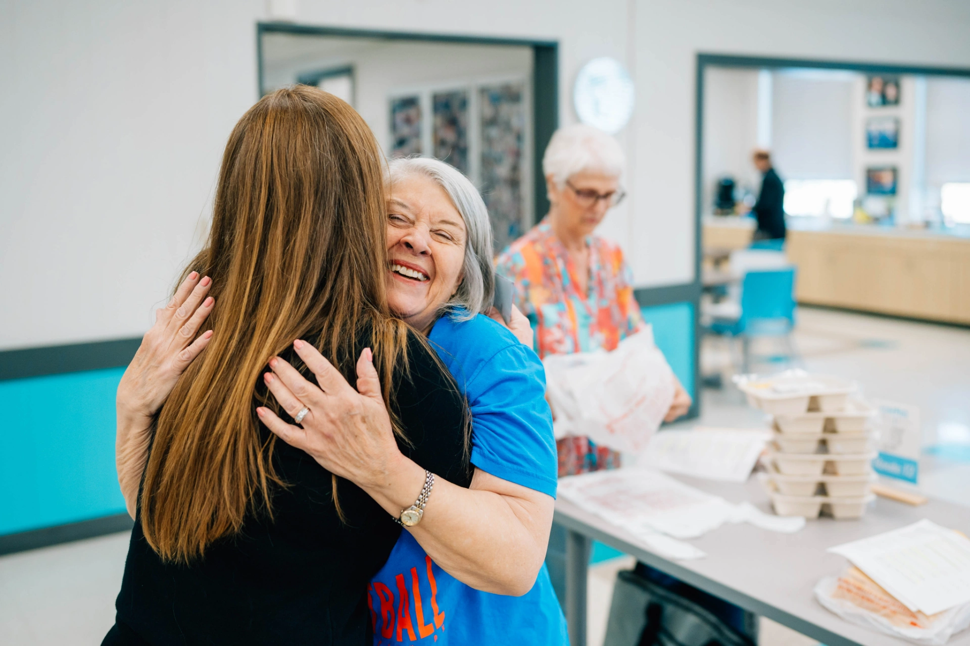 A smiling older woman hugs a younger woman in a bright community room, with another woman standing near a table stacked with food containers.