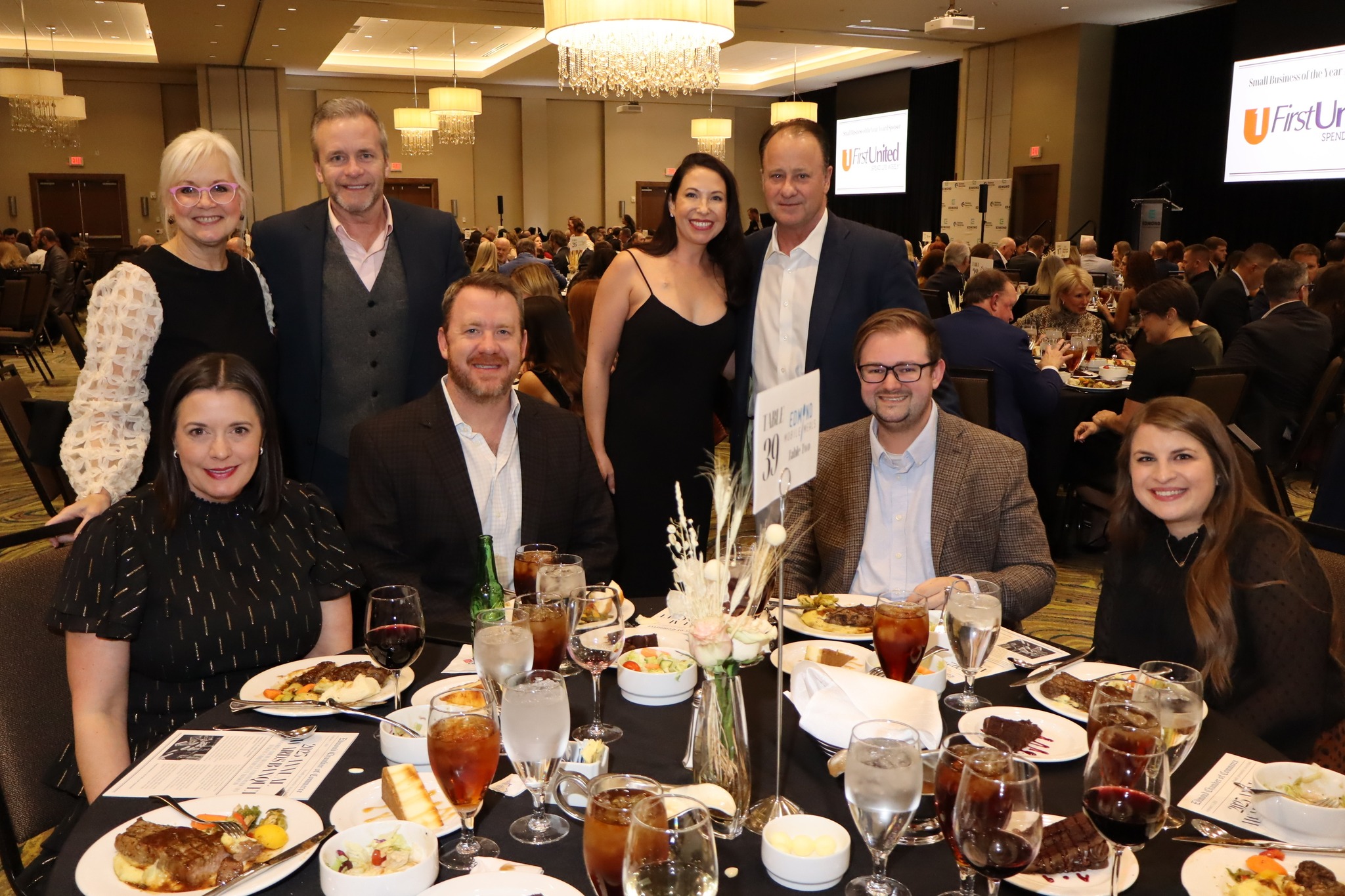 Eight people, four seated and four standing behind, gather smiling around a round dining table with plates of food, glasses of water, wine, and iced tea, in a large banquet hall with many other guests and chandeliers.