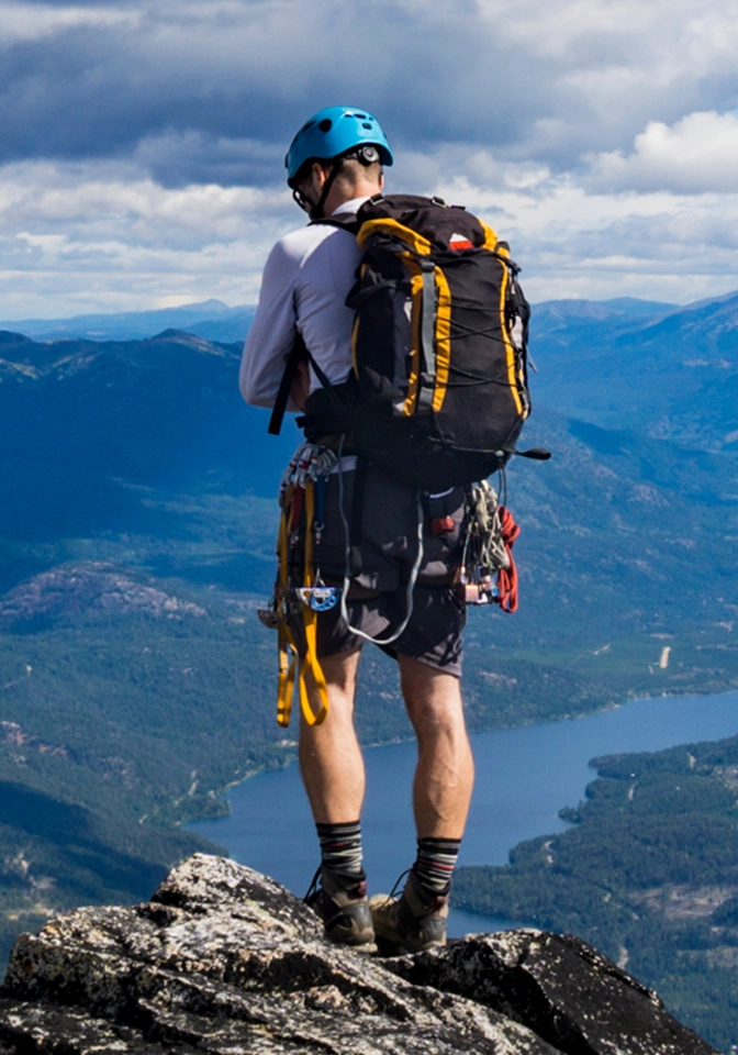 Hiker wearing a blue helmet and carrying a large backpack stands on a rocky mountain peak overlooking a lake and forested valleys below.