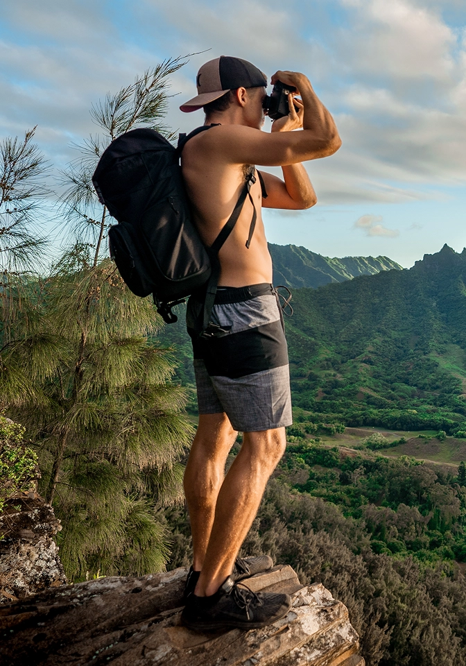 Shirtless man wearing a backward cap and backpack taking a photo with a camera atop a rocky cliff overlooking lush green mountains under a partly cloudy sky.