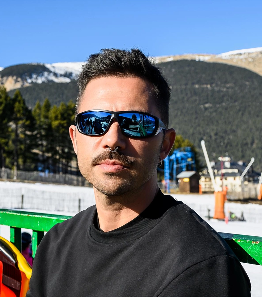 Man wearing blue mirrored sunglasses and a black sweatshirt with snowy mountains and trees in the background.