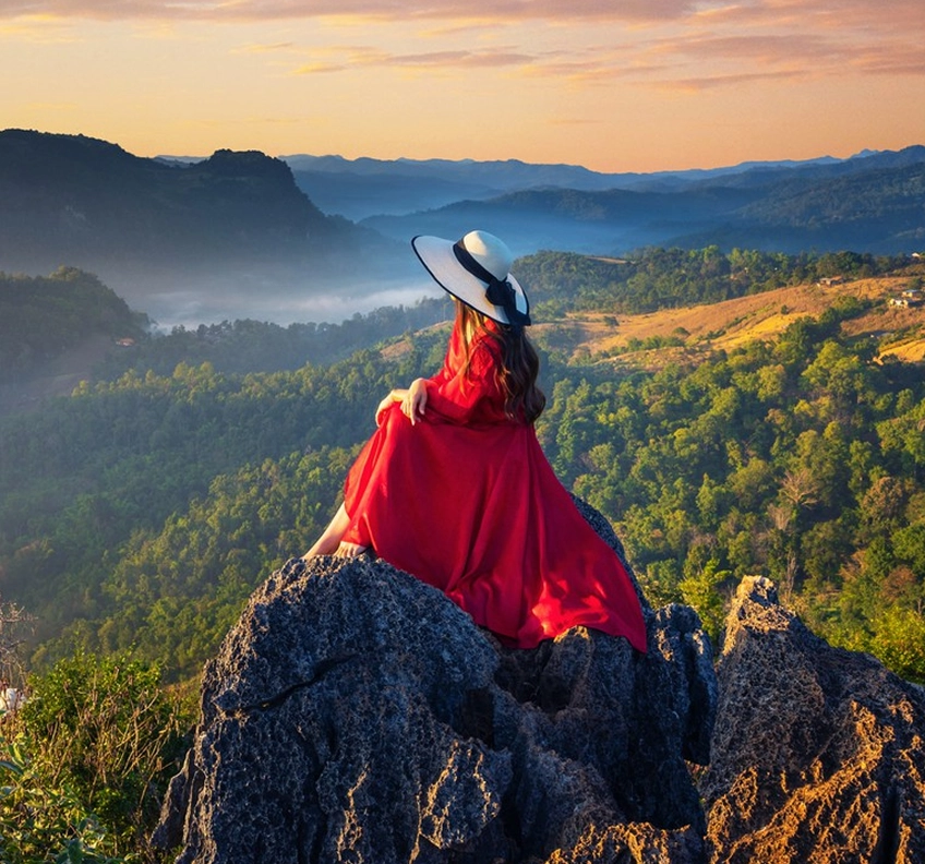 Woman in a red dress and wide-brimmed hat sitting on a rocky cliff overlooking a green valley at sunset.