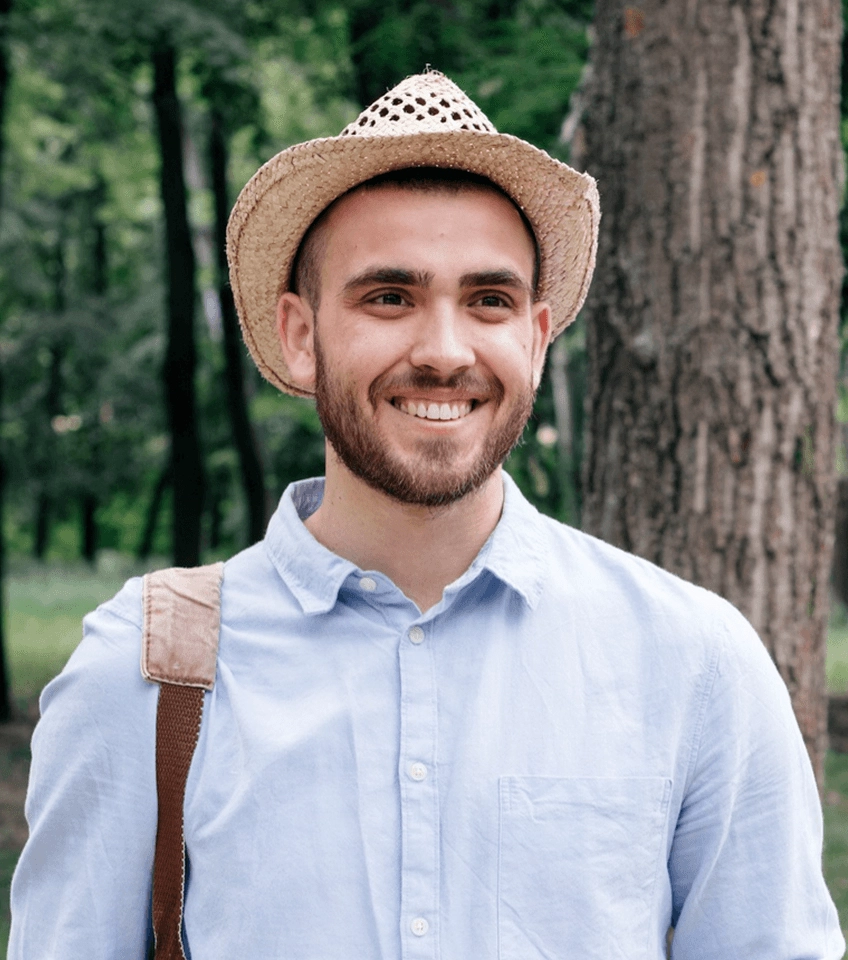 Smiling man wearing a straw hat and light blue shirt with a backpack strap over one shoulder standing outdoors near trees.