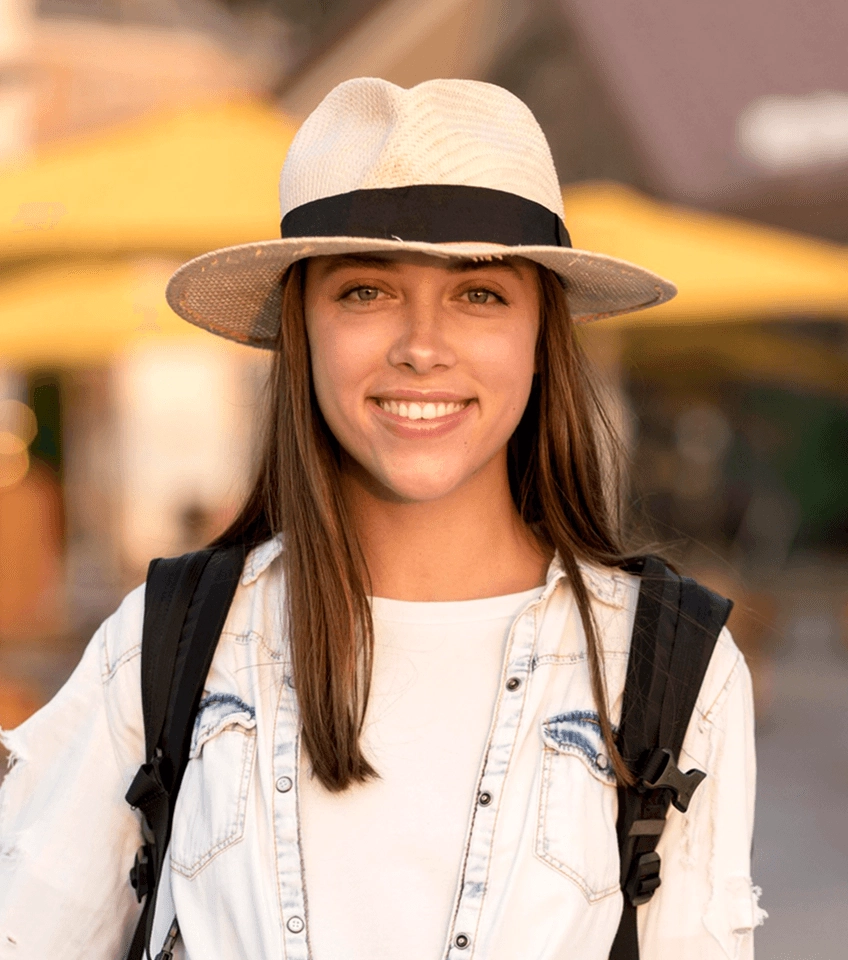 Smiling young woman wearing a white hat and denim jacket with backpack outdoors.