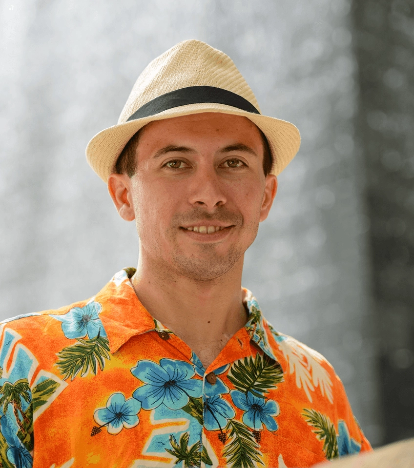 Man smiling wearing a beige fedora and orange Hawaiian shirt with blue flowers and green leaves.