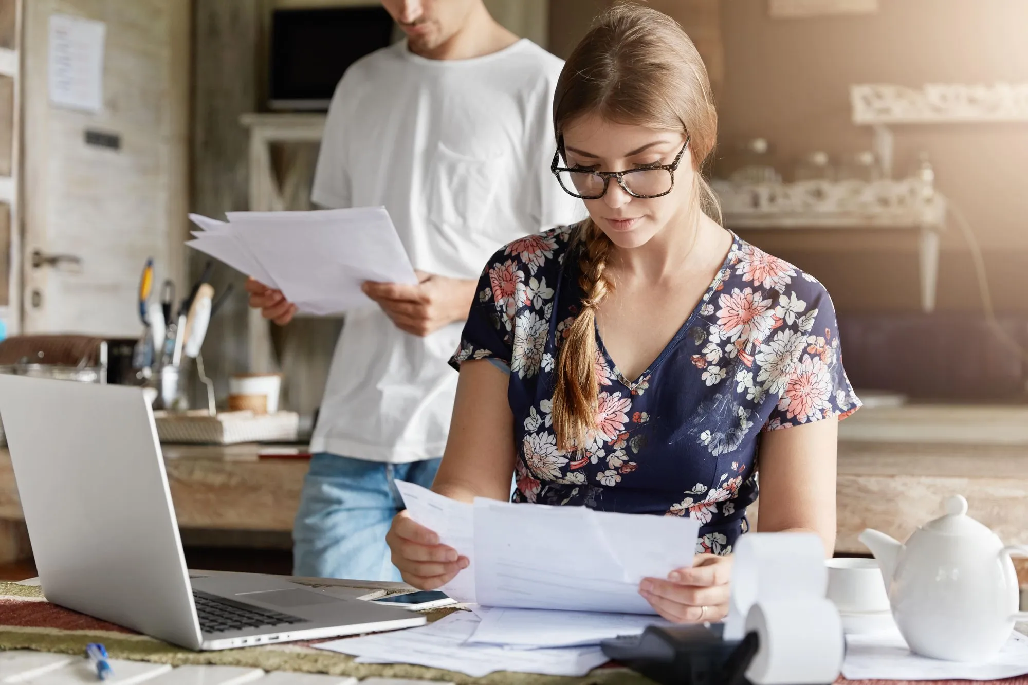 Woman with glasses reviewing papers at a table with laptop, man in background reading documents.
