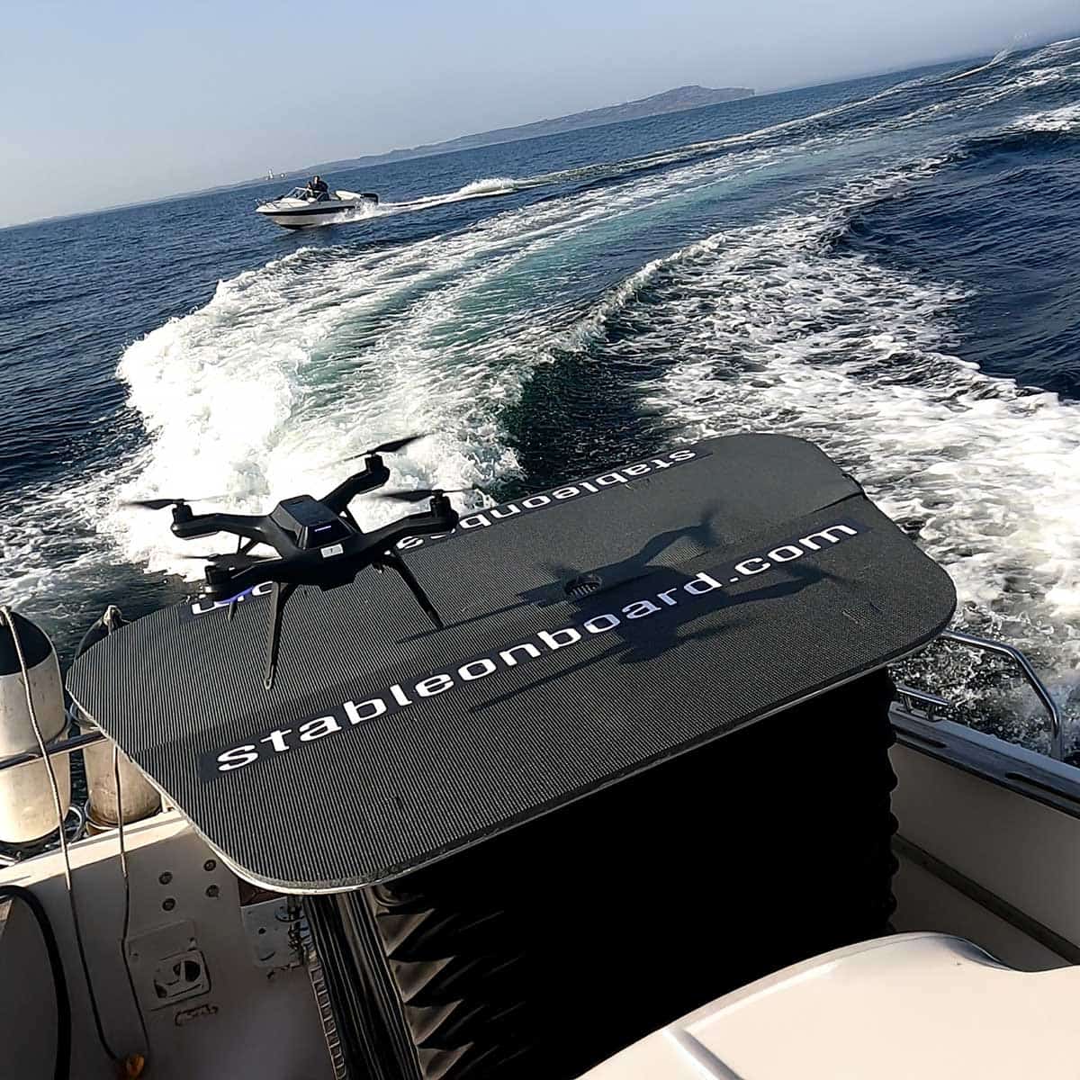 A black drone hovers over a platform labeled Stableonboard.com on a boat at sea for stable landing
