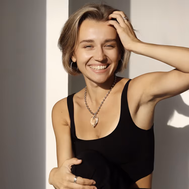 Smiling young woman with short hair wearing a black tank top and heart-shaped necklace in natural light.