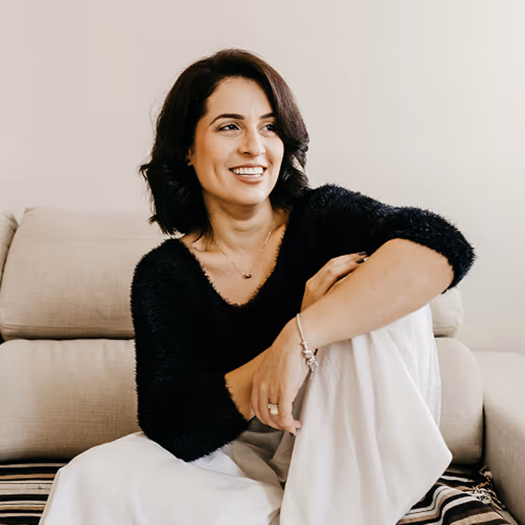Woman with dark hair wearing a black sweater and white pants sitting on a couch, smiling and looking right.