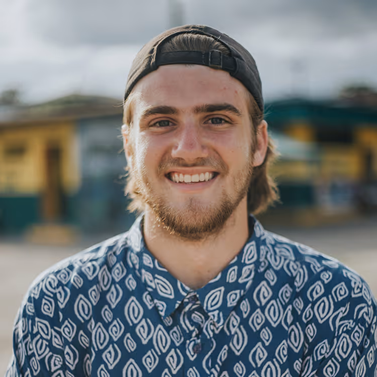 Smiling young man with beard in patterned shirt and backward cap outdoors with blurred background.