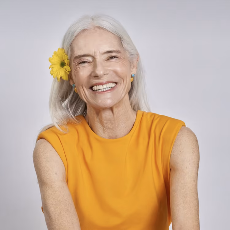 Smiling elderly woman with gray hair, wearing a sleeveless orange top and a yellow flower in her hair.