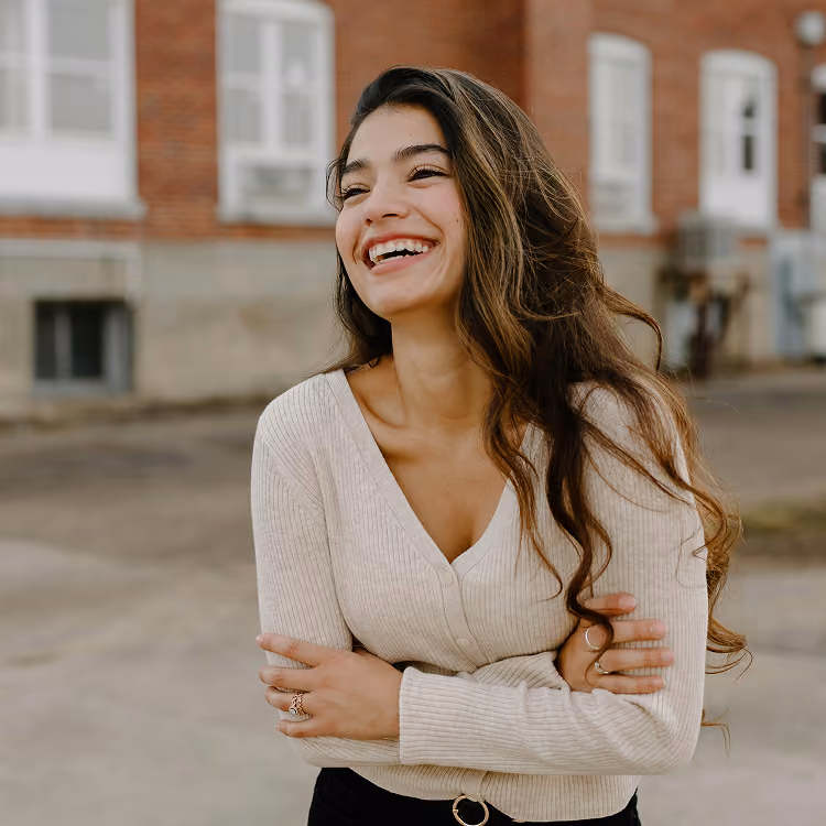 Smiling woman with long hair in a beige sweater standing outside in front of a brick building.