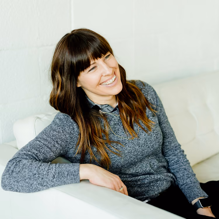 Smiling woman with brown hair sitting on a white couch wearing a gray sweater and watch.