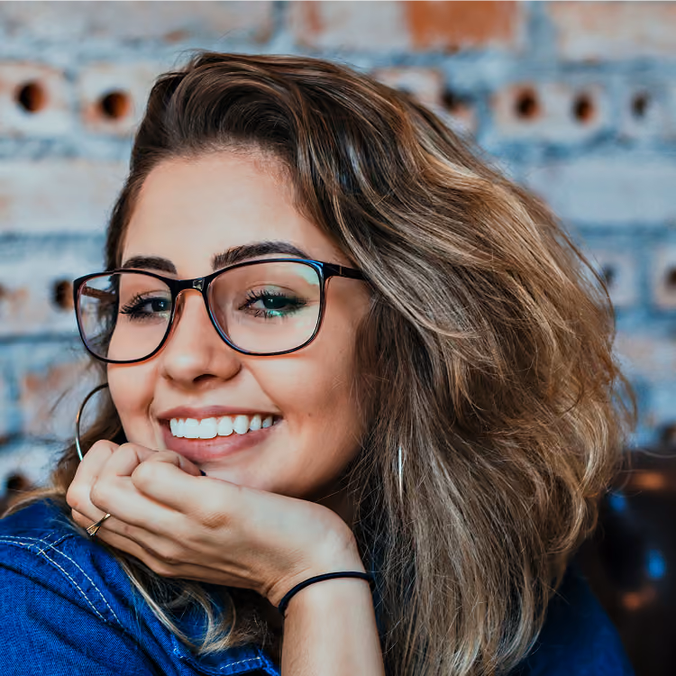 Smiling woman with wavy hair wearing glasses and resting her chin on her hand against a brick wall.