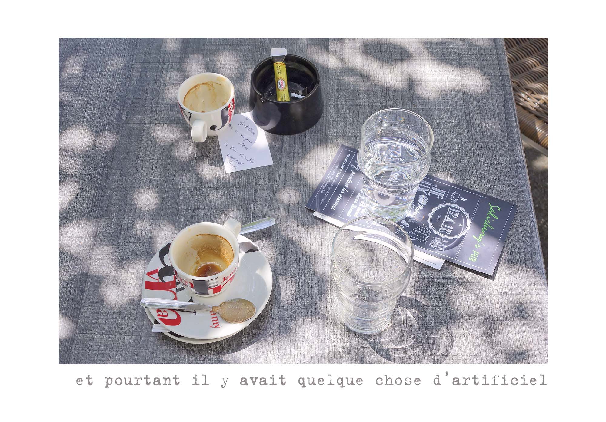Deux tasses à café vides posées sur une table grise avec deux verres d'eau, un cendrier noir contenant un sucre en sachet, une note manuscrite et un menu de bar au style vintage, avec des ombres d'arbres qui tombent sur la table.