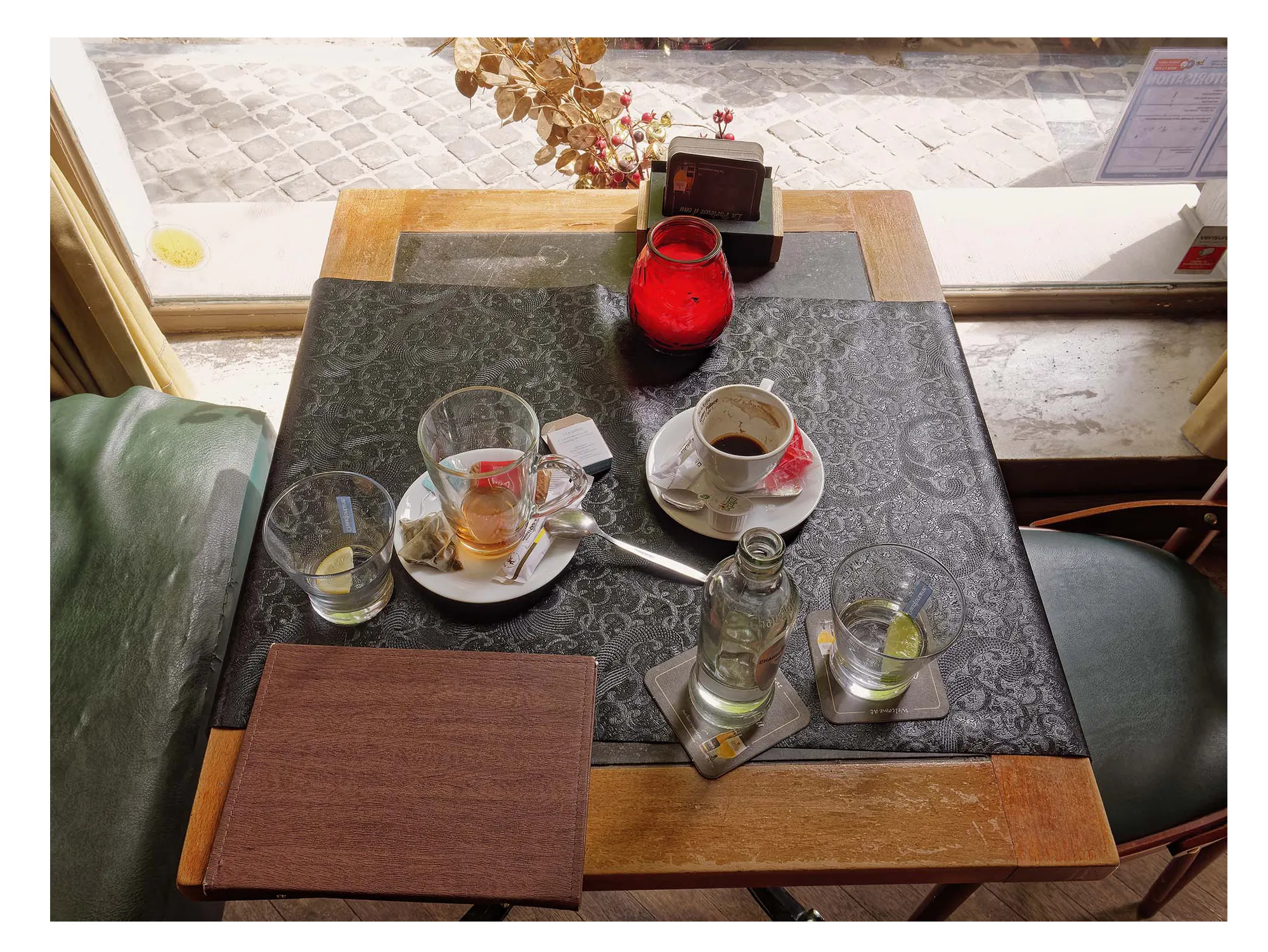 Table near window with cups, glasses with lemon, bottle, red candle, and brown notebook.