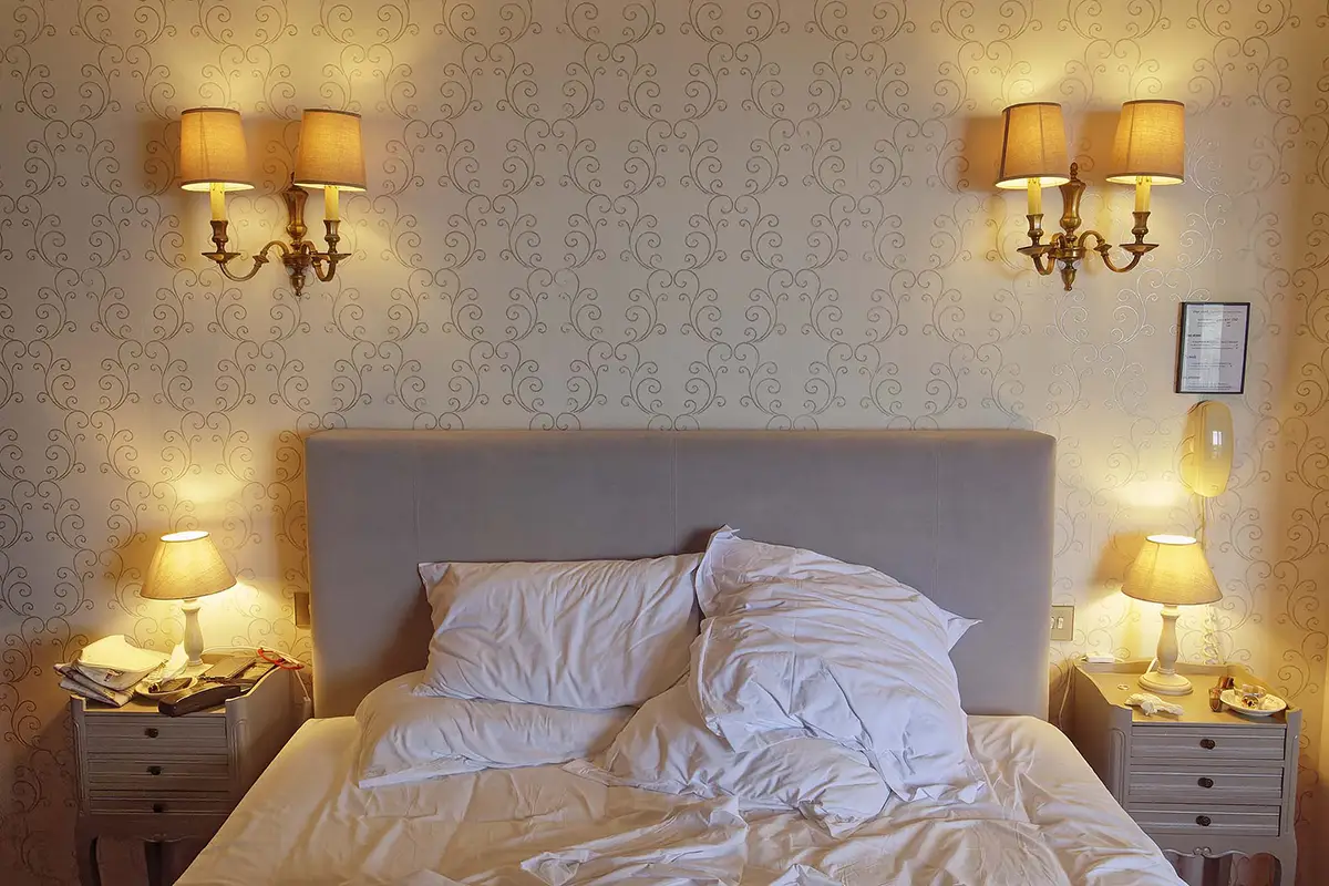 Lit double avec tête de lit beige, deux oreillers et une couette froissée, deux tables de chevet avec lampes allumées, appliques murales et papier peint décoratif derrière.