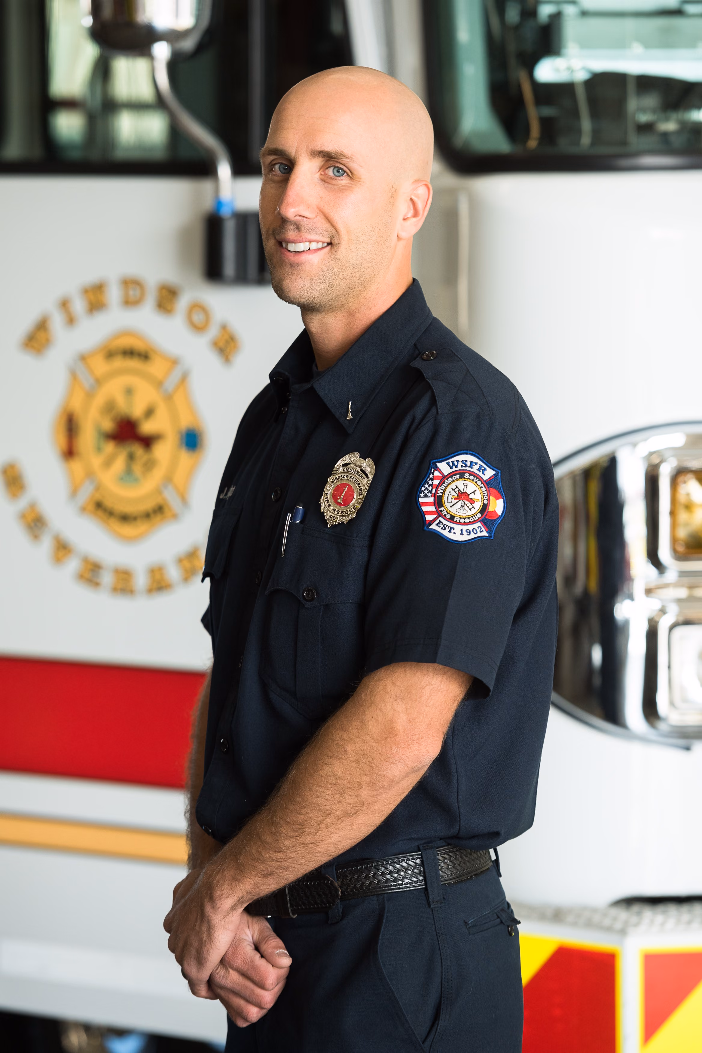 Smiling firefighter with badge and patch stands in front of a fire truck labeled Windsor Severance.