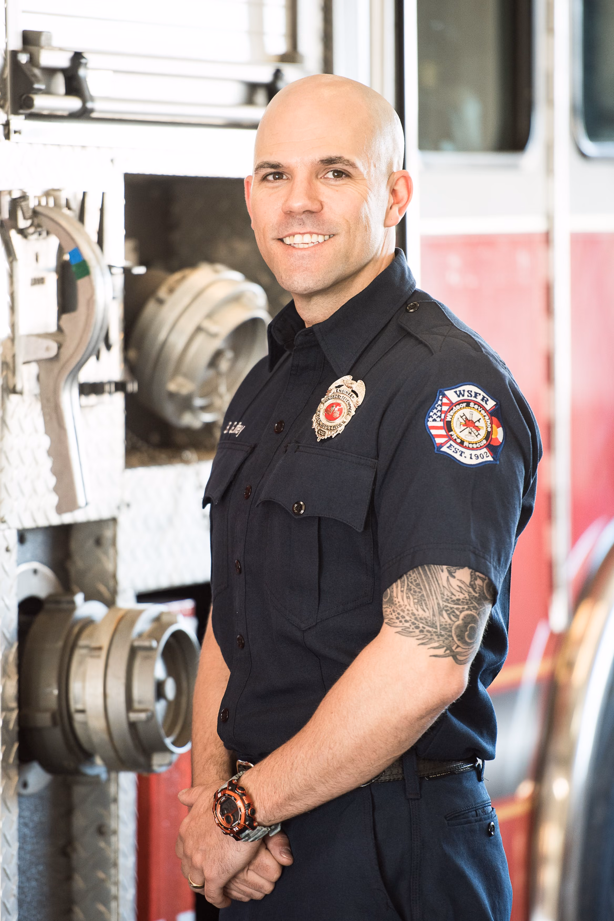 Smiling firefighter in uniform with tattooed arm standing by a fire truck with hose connectors.