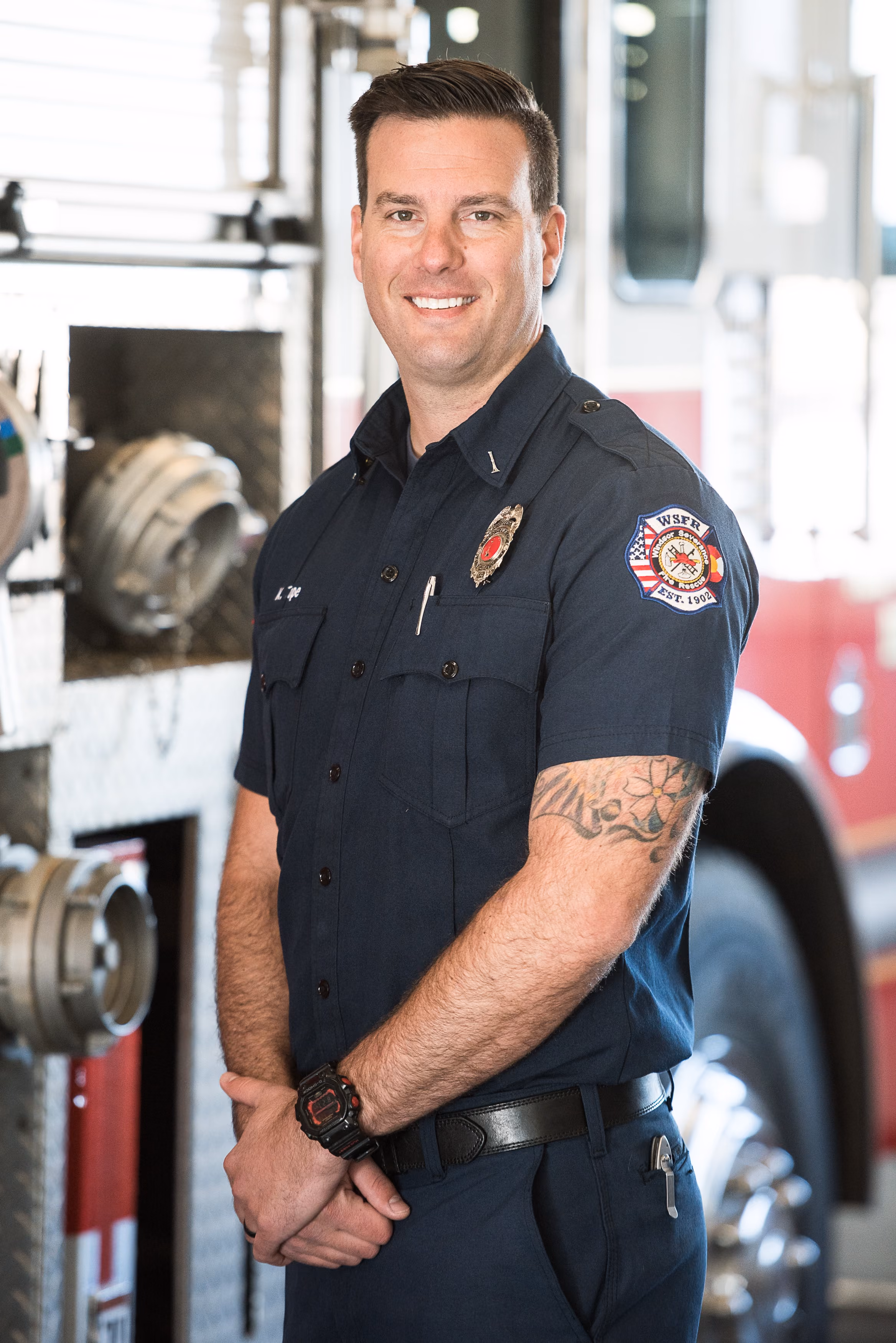 Smiling firefighter in uniform with tattooed arm standing in front of fire truck.