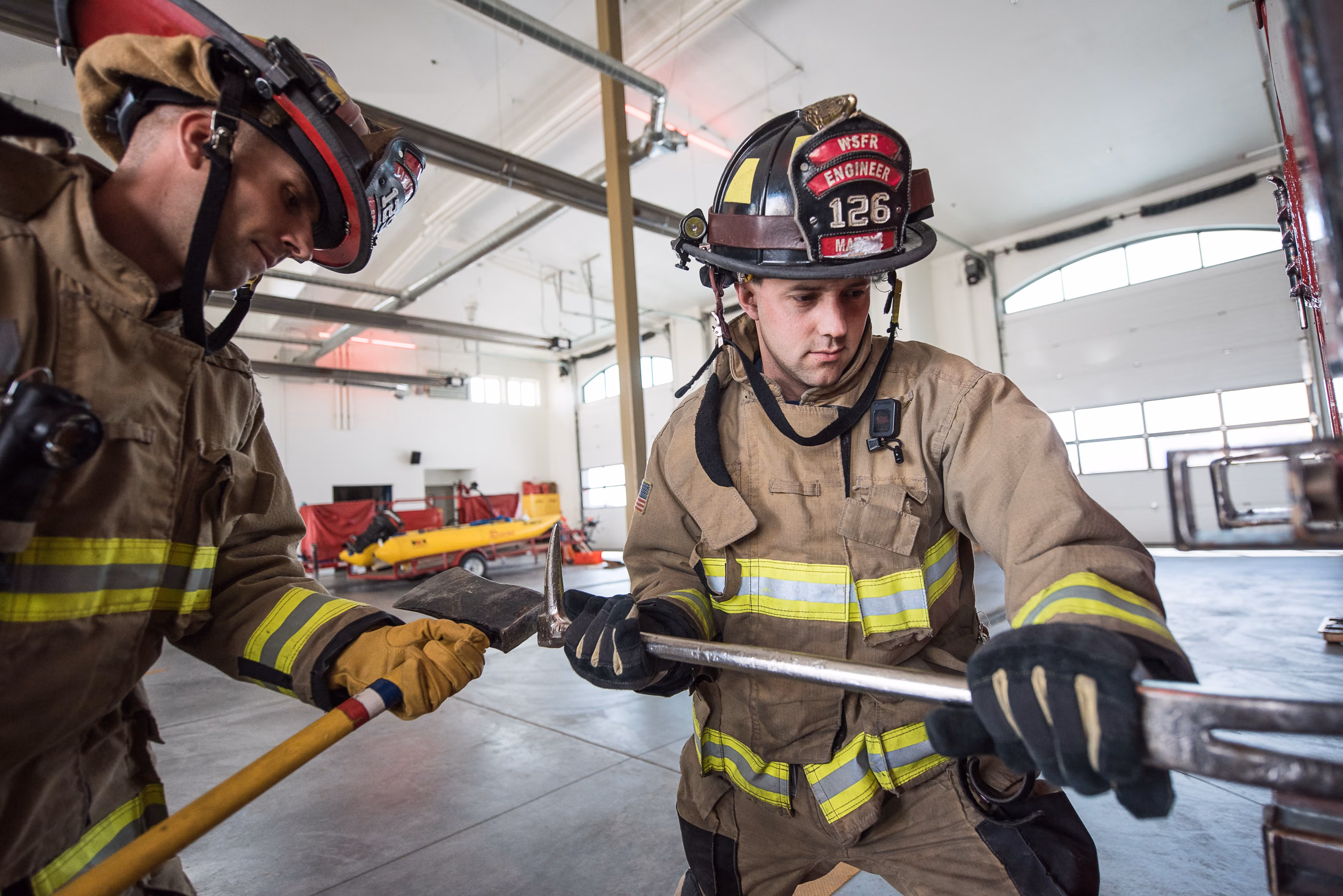 Two firefighters in gear using tools inside a fire station with large garage doors.