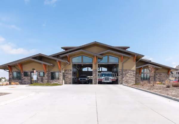Fire station building with two open garage bays and fire trucks inside on a clear day.