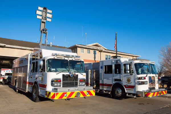 Two white fire trucks with red stripes parked outside a fire station under a clear blue sky.