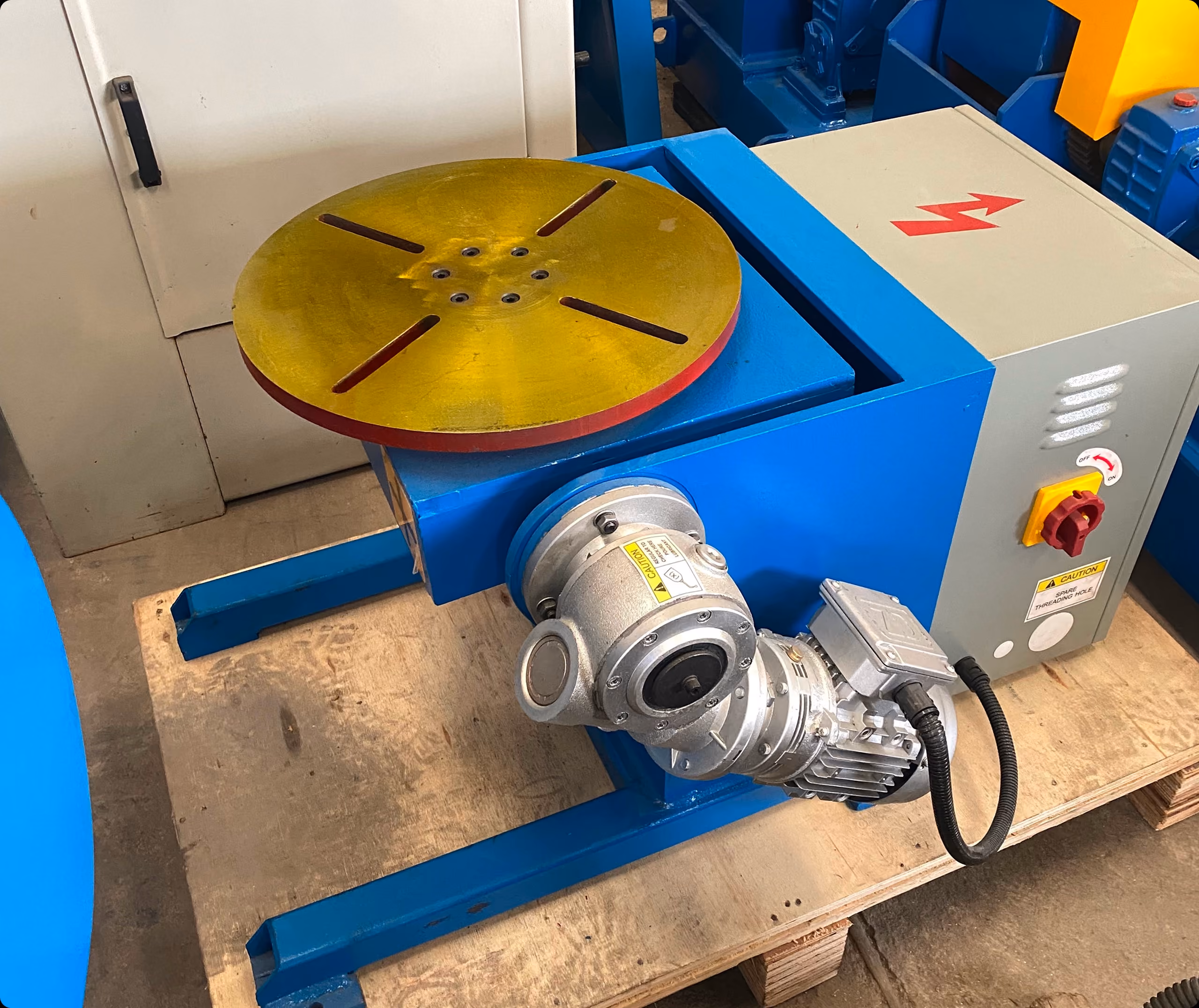 Blue industrial rotary table machine with yellow circular platform and attached motor on wooden floor.
