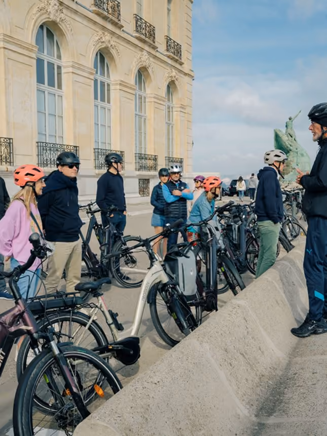 Un groupe de cyclistes portant des casques écoutent un guide près d'un bâtiment historique.