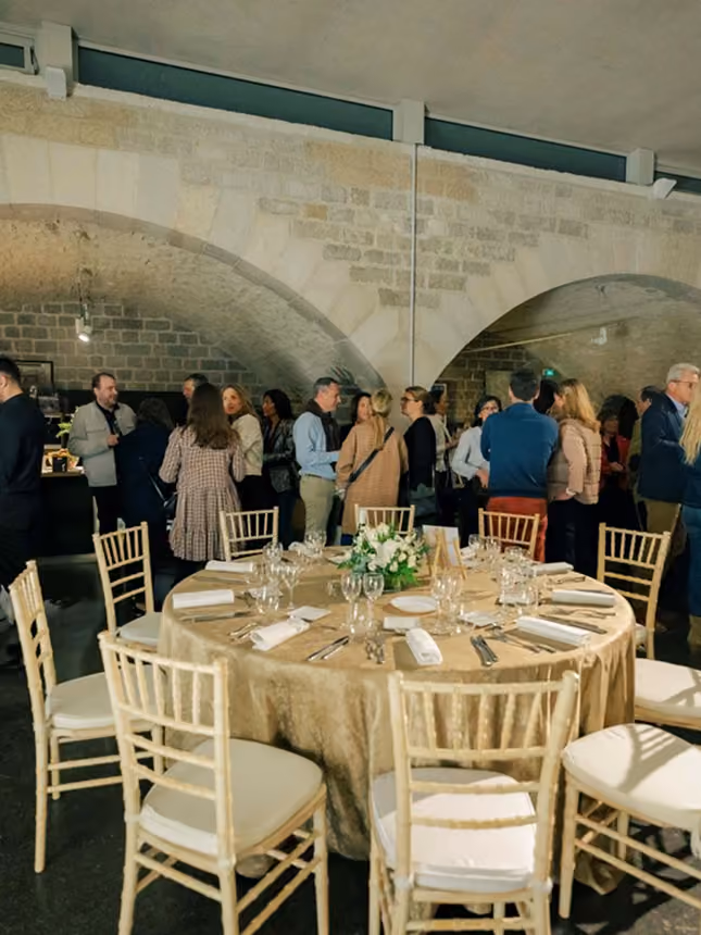 Salle avec une grande table dressée et des invités discutant sous des arches en pierre.