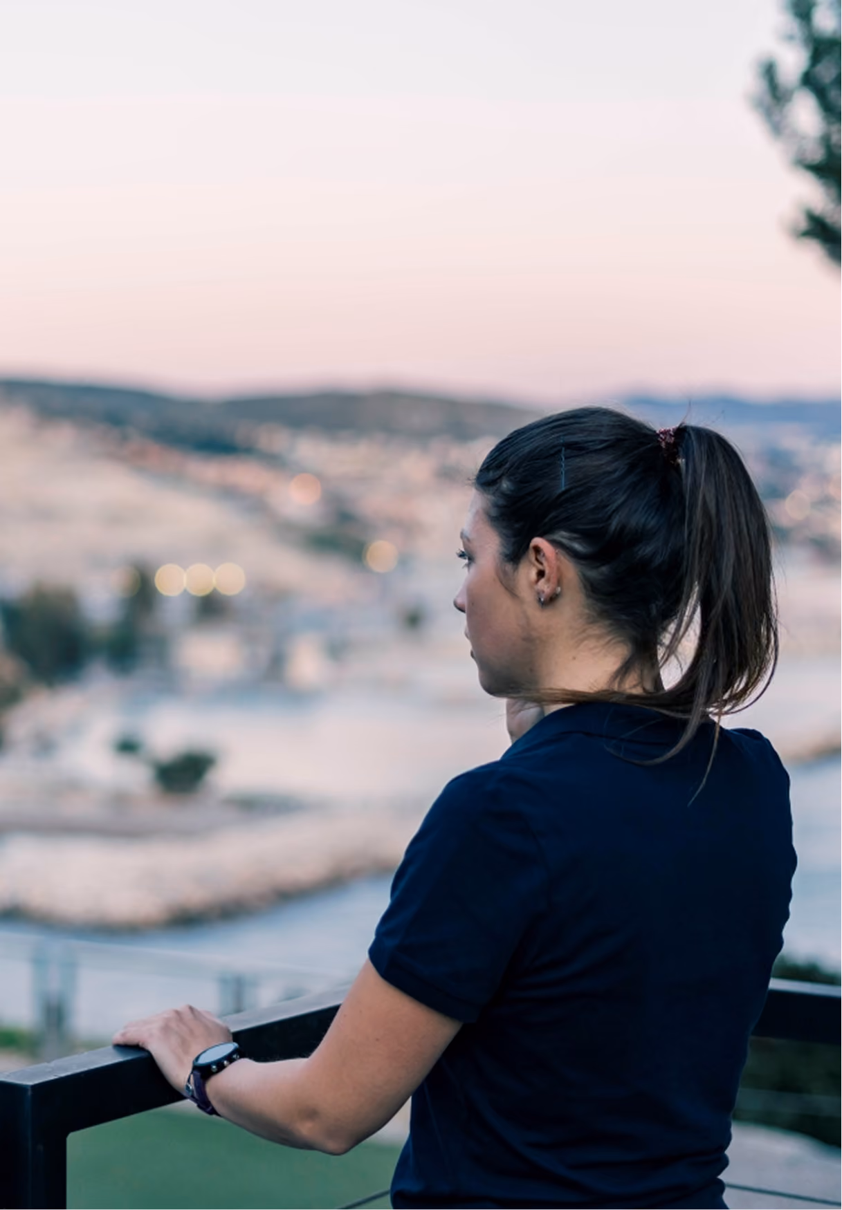 Femme vue de dos appuyée sur une rambarde, regardant un paysage urbain flou au crépuscule.