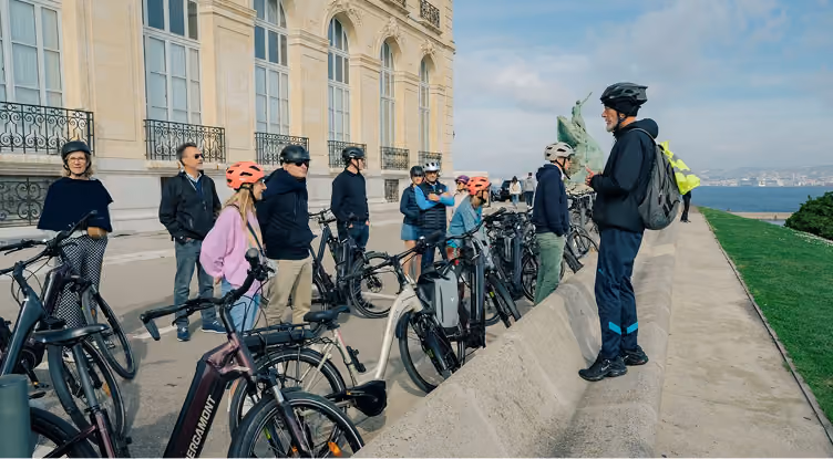 Groupe en tenue de vélo avec casques, écoutant un guide près d'un bâtiment et statue au bord de l'eau.
