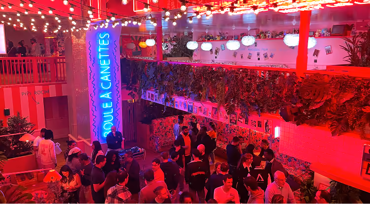 Vue intérieure d'une fête animée avec des lumières colorées, des invités et une grande enseigne bleue lumineuse.