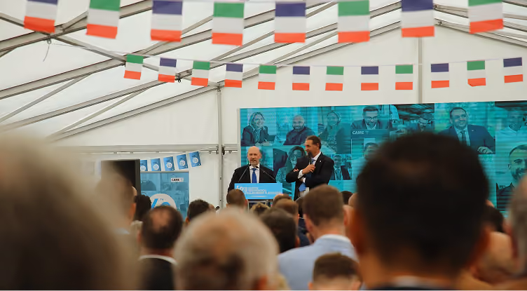 Deux hommes parlant devant un podium lors d'un événement sous tente décorée de drapeaux.