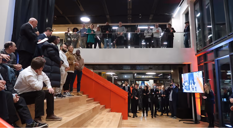 Un groupe de personnes écoute un orateur parlant au micro dans un espace contemporain avec escaliers et écran.