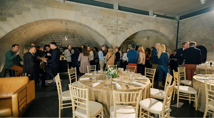 Un groupe de personnes discutant dans une salle avec des tables rondes et des chaises sous des voûtes en pierre.