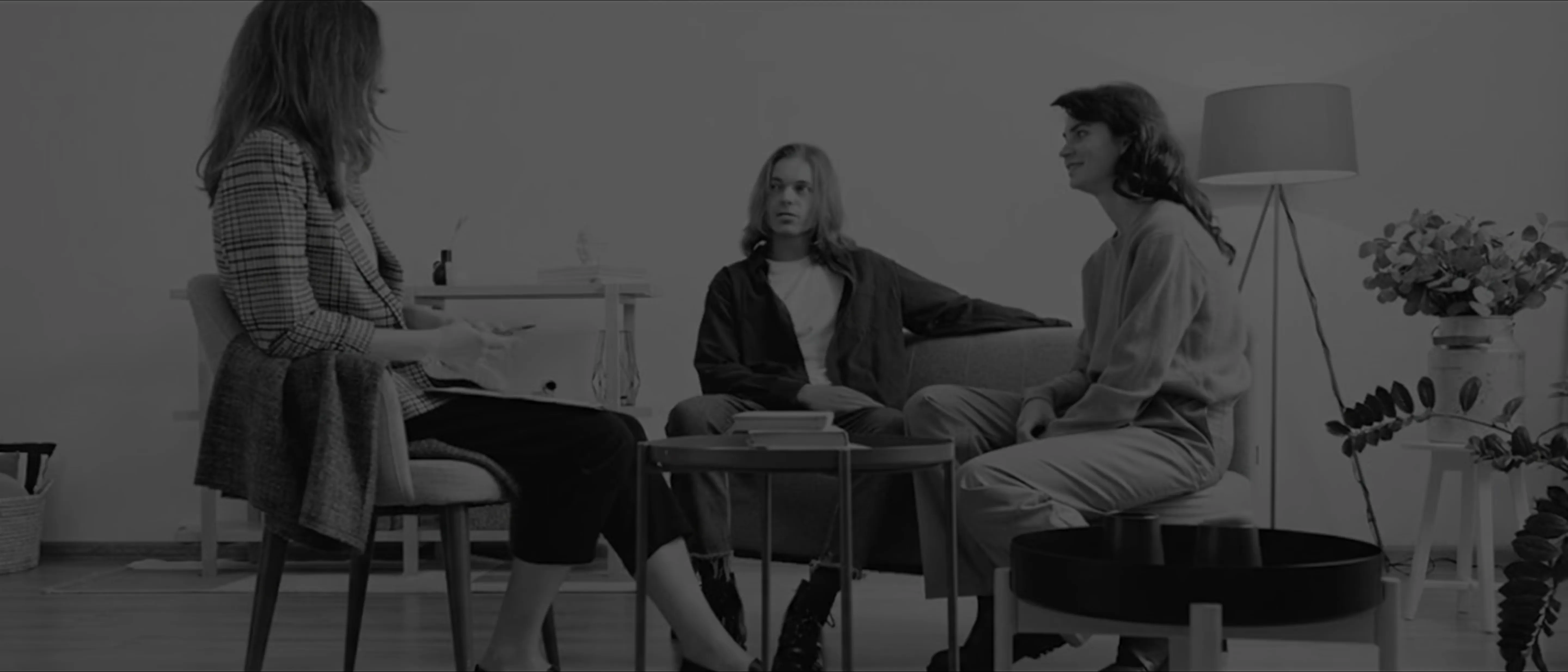 Three people seated in a modern living room having a conversation, with a lamp and plant in the background.