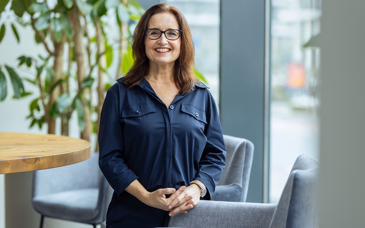Smiling woman with glasses in navy blue shirt standing by table and chairs near window indoors.
