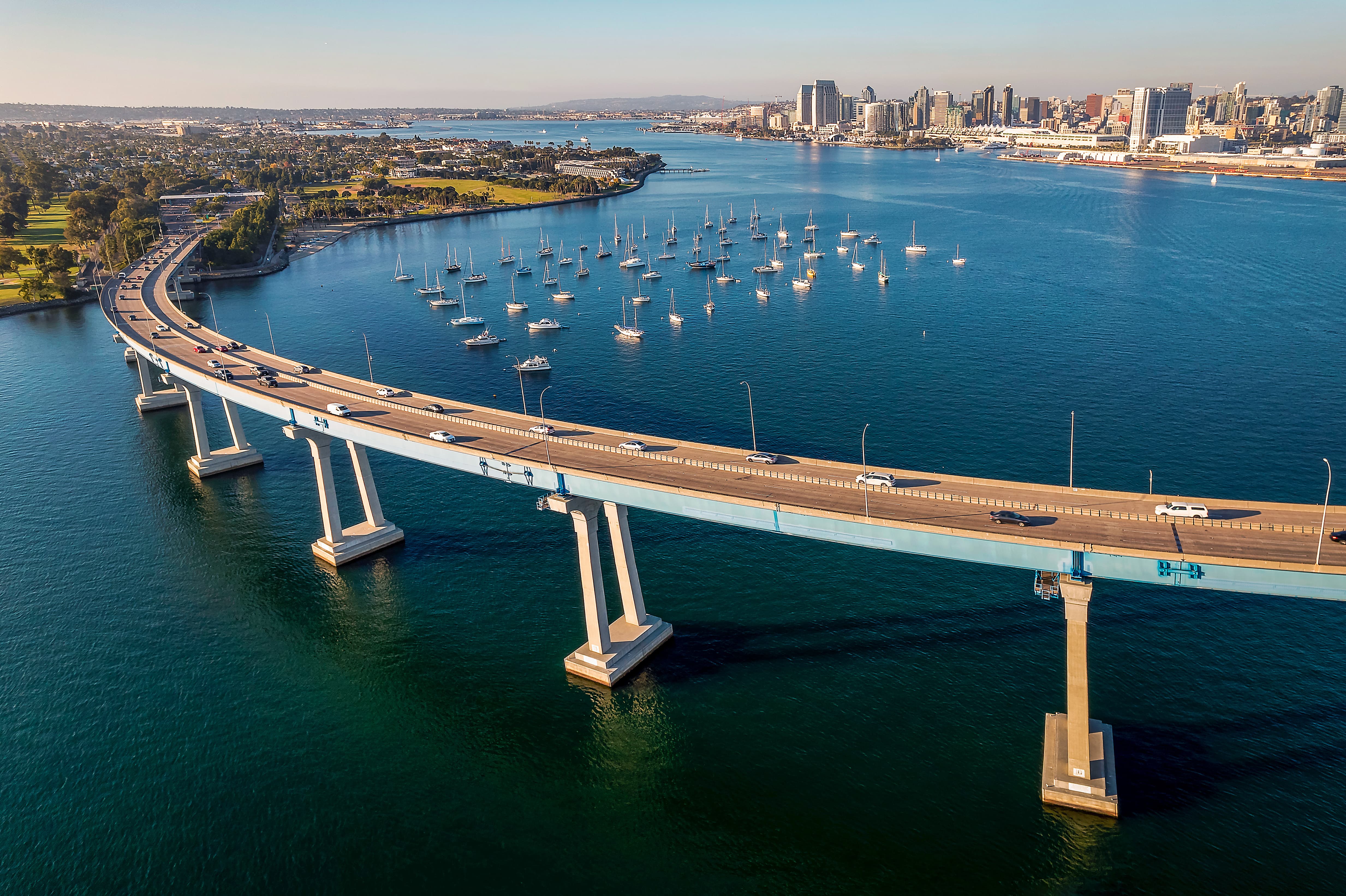 Aerial view of a curved bridge over blue water with sailed boats and a city skyline in the background.