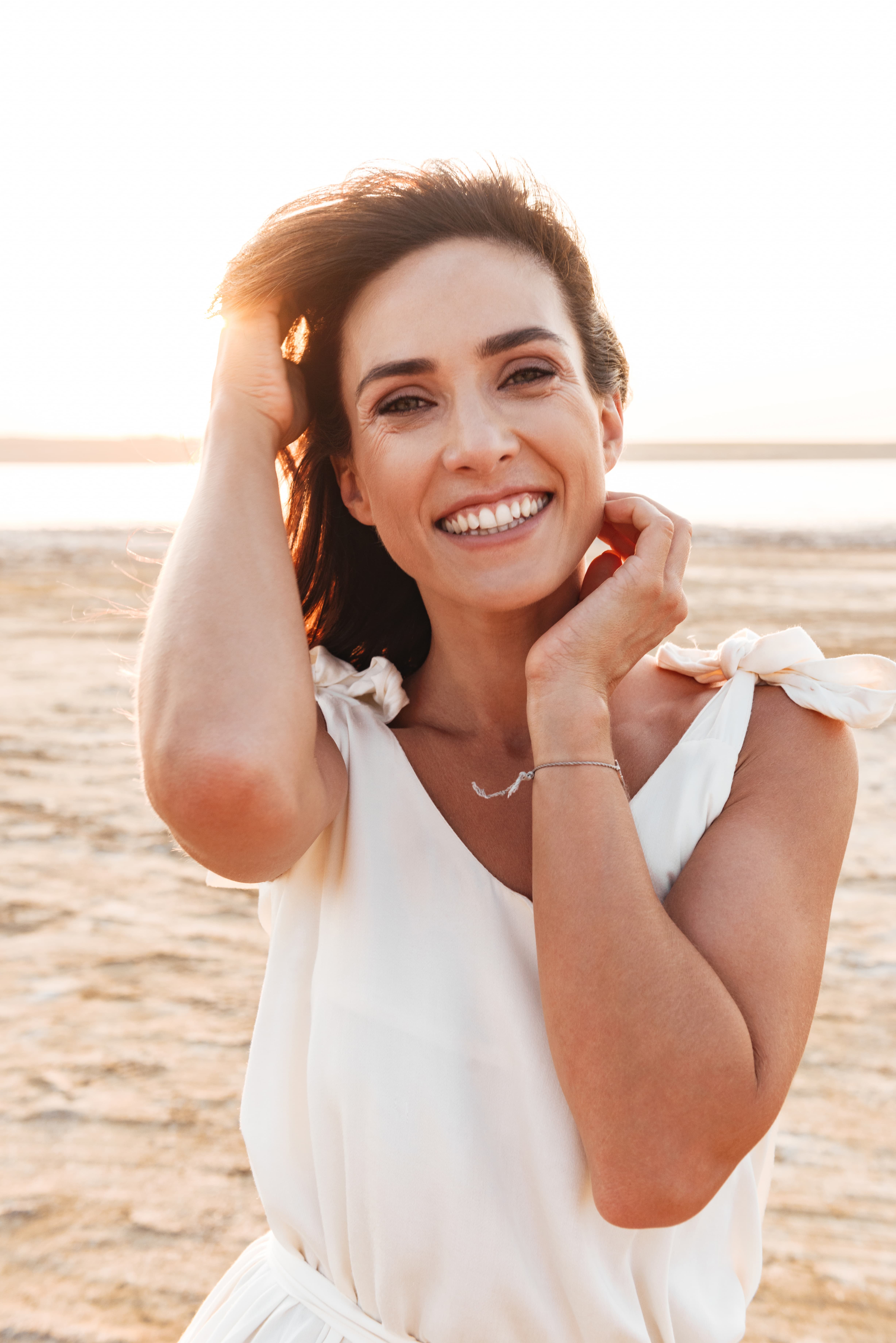 Smiling woman in white dress touching her hair outdoors with a sandy background at sunset.