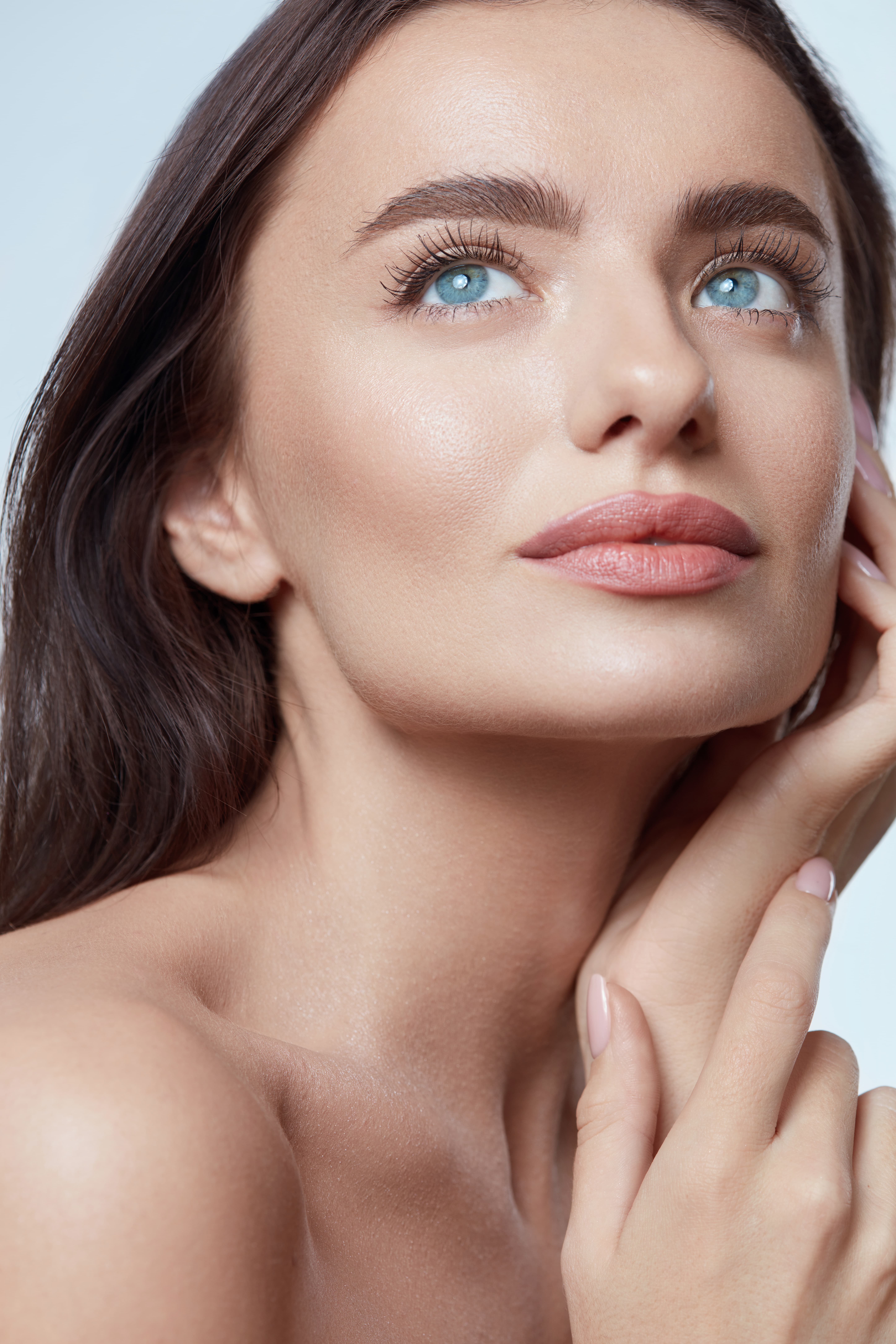 Close-up of woman with clear skin, blue eyes, and pink lips touching her face against a light background.