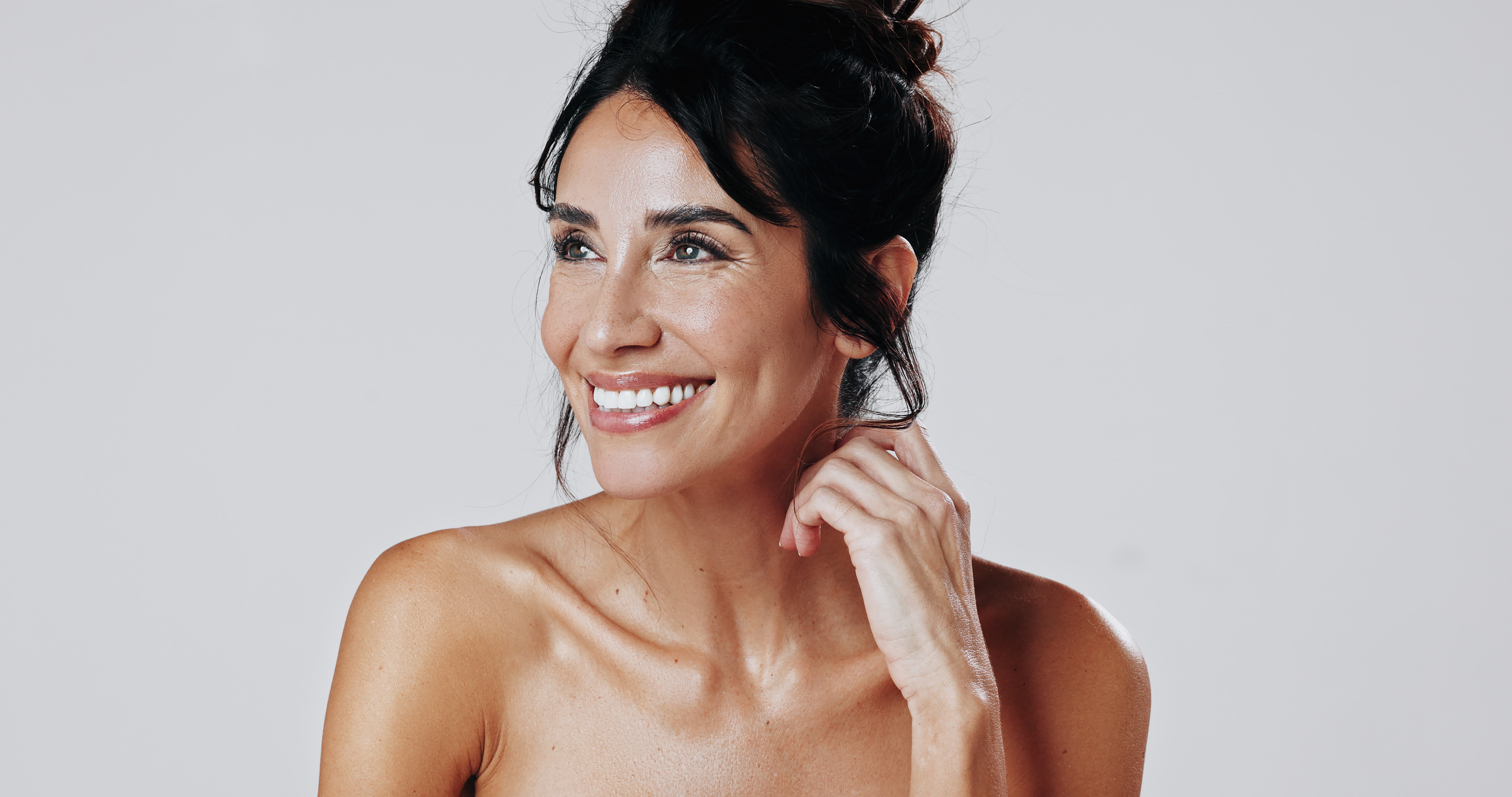 Smiling woman with dark hair in a bun looking to the side against a plain light gray background.