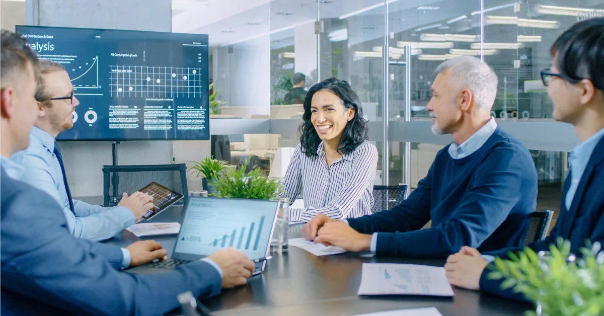 Smiling team at a meeting table with laptops and a tablet, a wall screen showing analysis charts